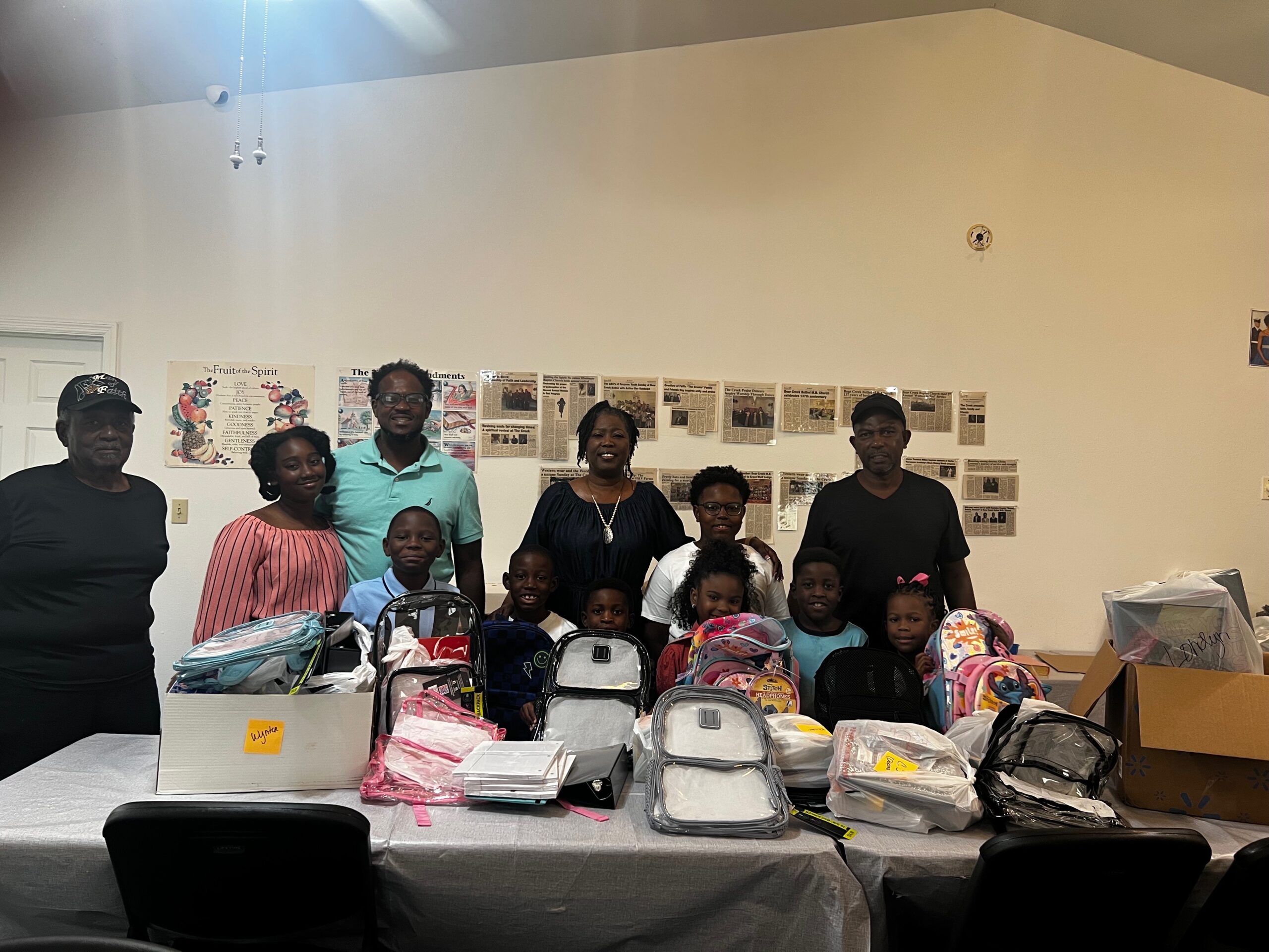 Group of adults and children pose behind a table filled with school supplies and backpacks in a room decorated with educational posters.