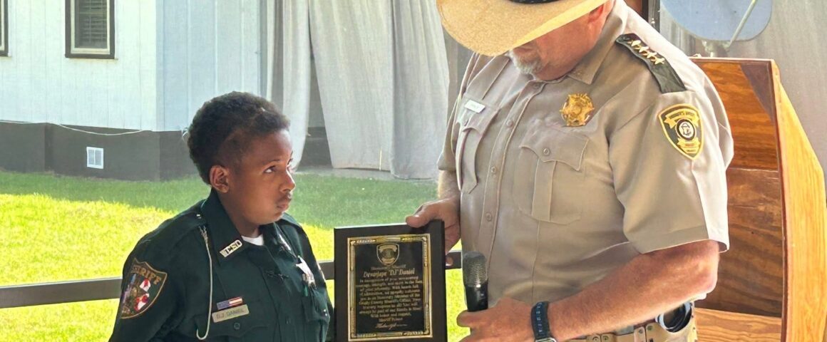 A uniformed sheriff presents a plaque to a young boy dressed in a sheriff’s uniform during a ceremony, standing together in a covered outdoor space.