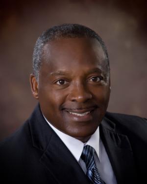 Professional headshot of Eugene Lamb in a dark suit and striped tie, smiling warmly against a softly lit studio background.