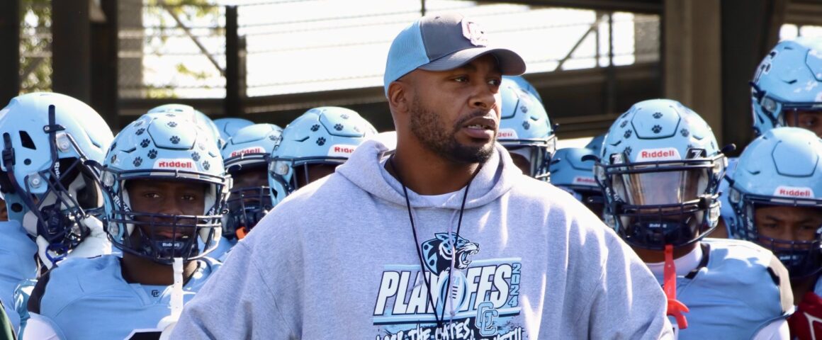 Embattled head coach Russell Ellington stands in front of his team, wearing a light blue and gray playoff sweatshirt, looking forward with determination as his players in full uniform prepare for the game behind him.