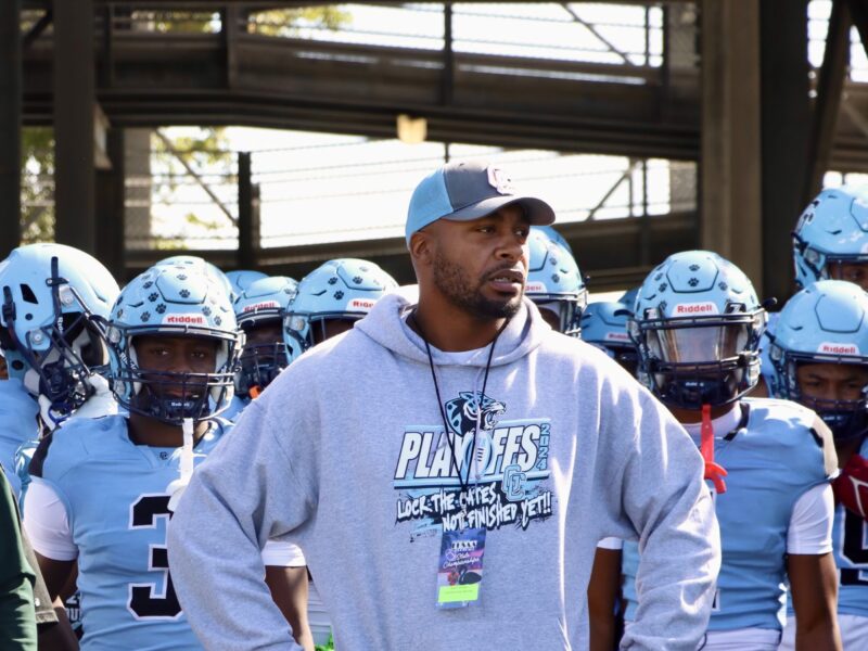 Embattled head coach Russell Ellington stands in front of his team, wearing a light blue and gray playoff sweatshirt, looking forward with determination as his players in full uniform prepare for the game behind him.