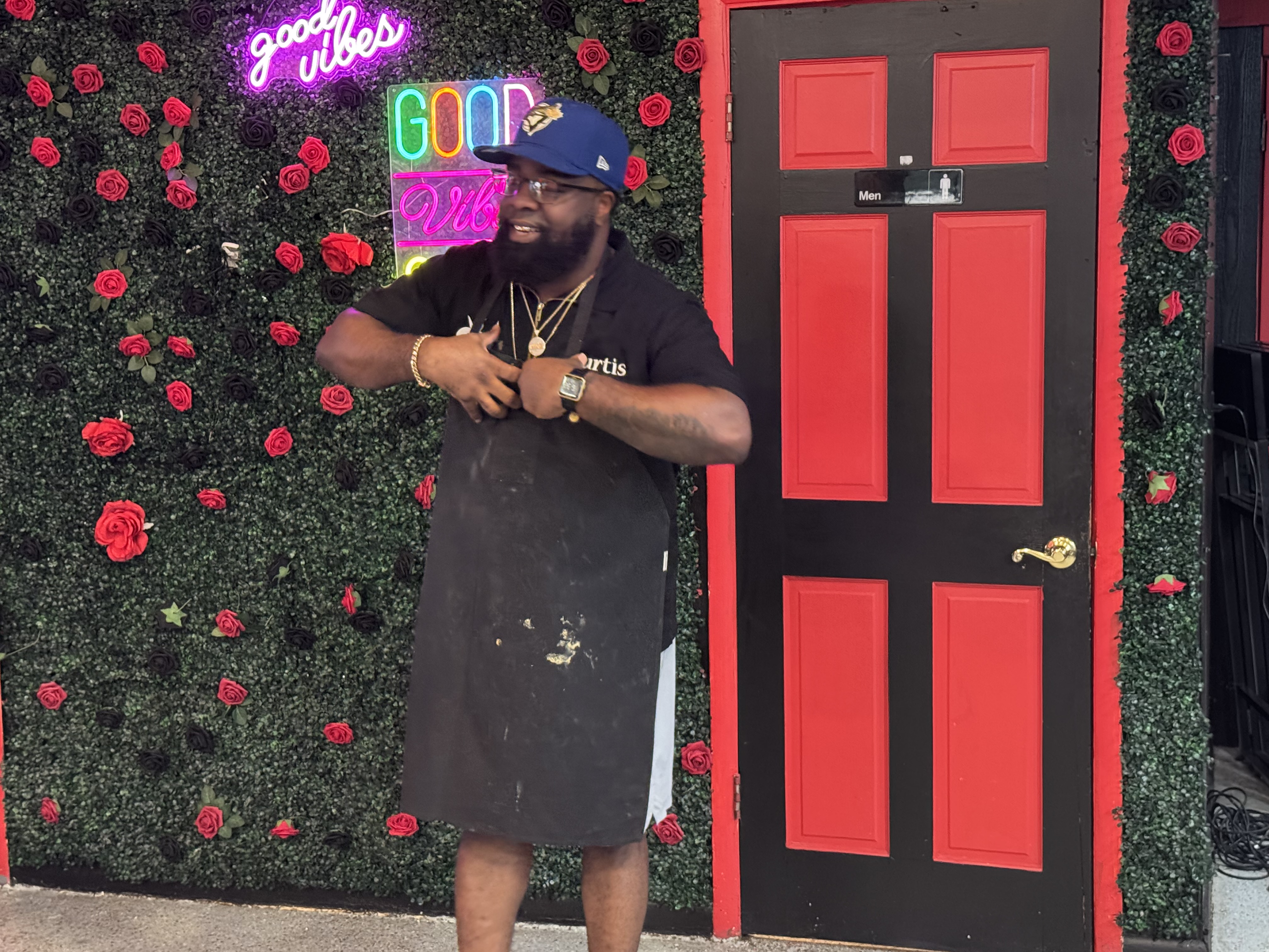 Man wearing an apron and gold jewelry stands smiling in front of a floral wall with neon "Good Vibes" signs and a red-and-black door.