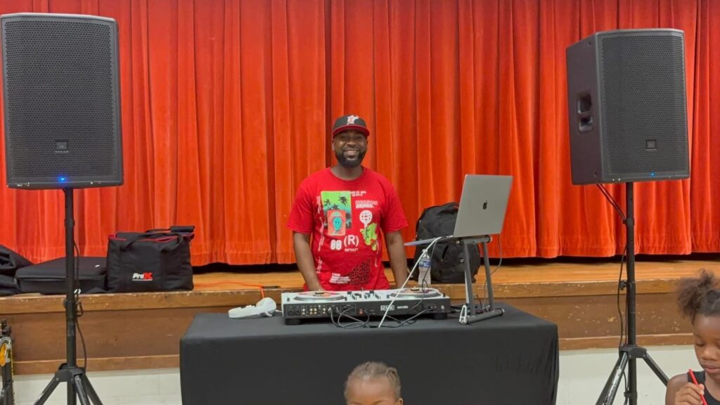 DJ stands behind a turntable with large speakers in front of a red stage curtain, smiling during a school or community event.