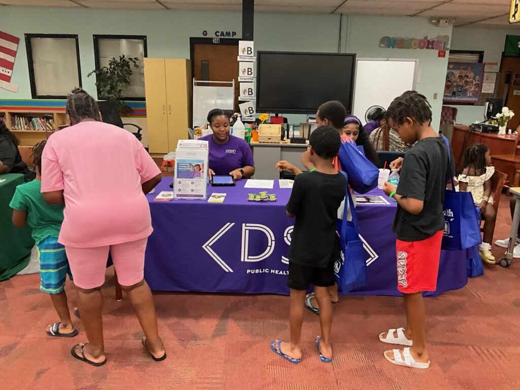 Children and adults gather around a purple outreach table staffed by representatives from the Department of Public Health inside a library or classroom.