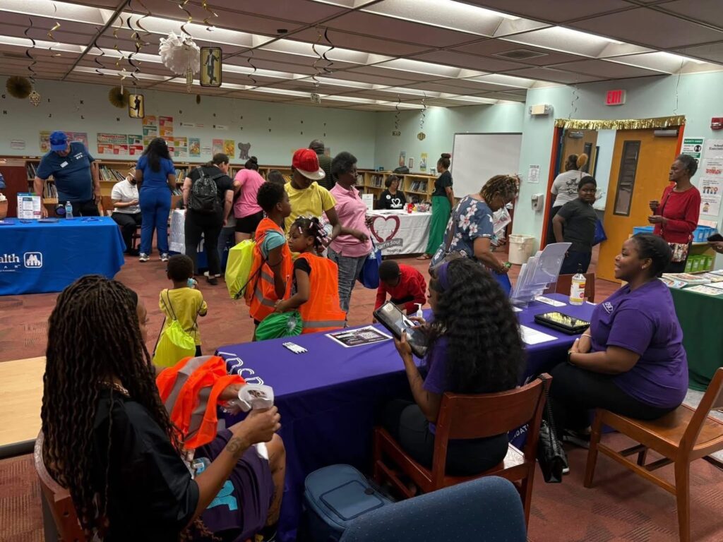 Indoor community event with families visiting resource tables staffed by local organizations; children are seen with drawstring bags and snacks.