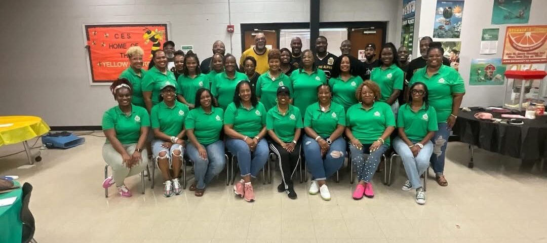 Large group of adults pose together in matching green shirts for a group photo at an indoor school or community event.