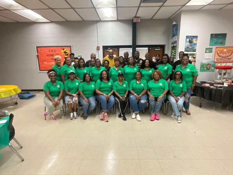 Large group of adults pose together in matching green shirts for a group photo at an indoor school or community event.