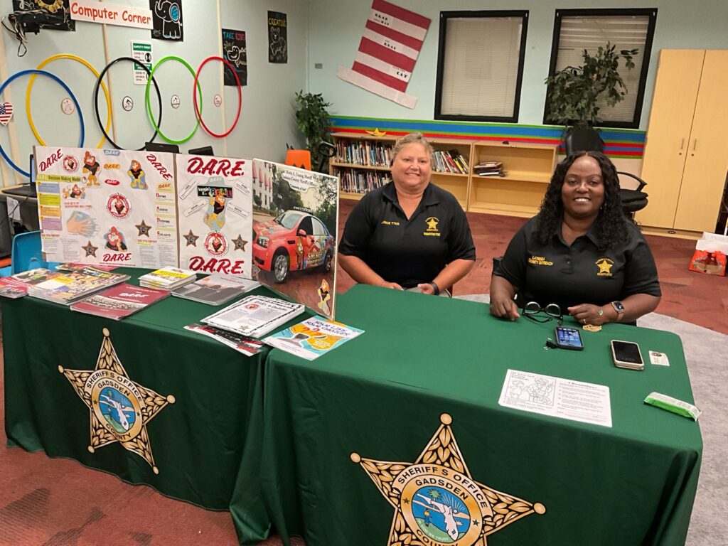 Two women from the Gadsden County Sheriff's Office sit behind a green D.A.R.E. information table in a colorful library or learning center.