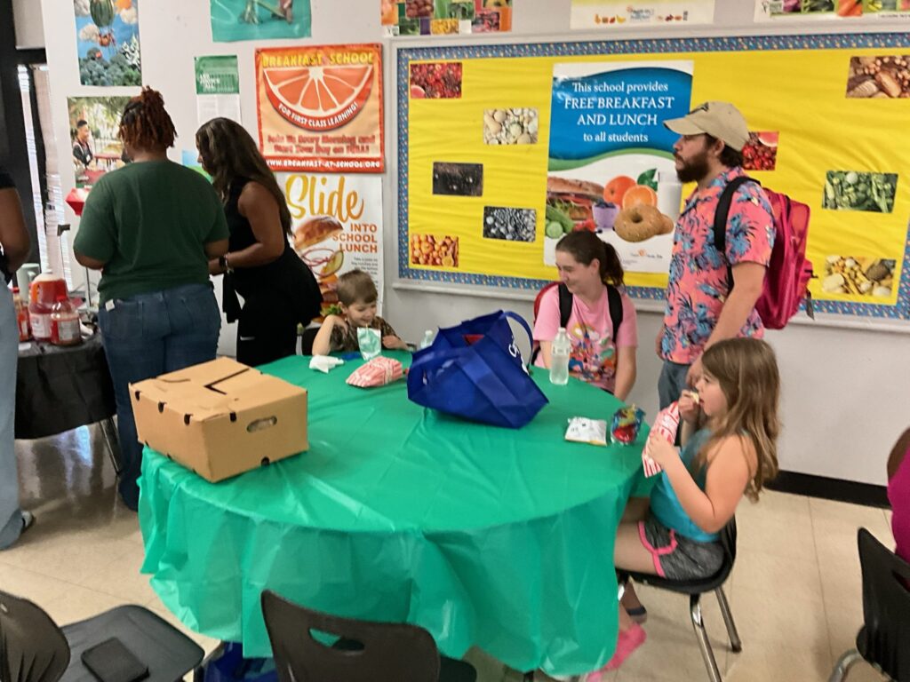 Children and adults sit and stand around a green table at a school event with posters promoting school meals and healthy eating on the wall.