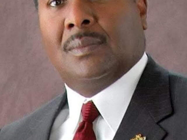 Formal portrait of Reginald James wearing a dark pinstripe suit, white shirt, and red tie, looking confidently at the camera with a neutral background.