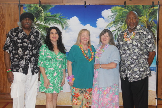 Five people in tropical shirts and leis pose together in front of a beach backdrop.