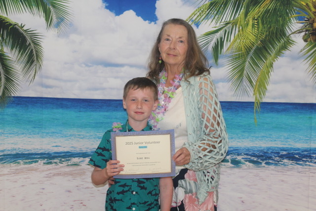 Woman and young boy pose with a “Junior Volunteer” certificate in front of a beach backdrop.