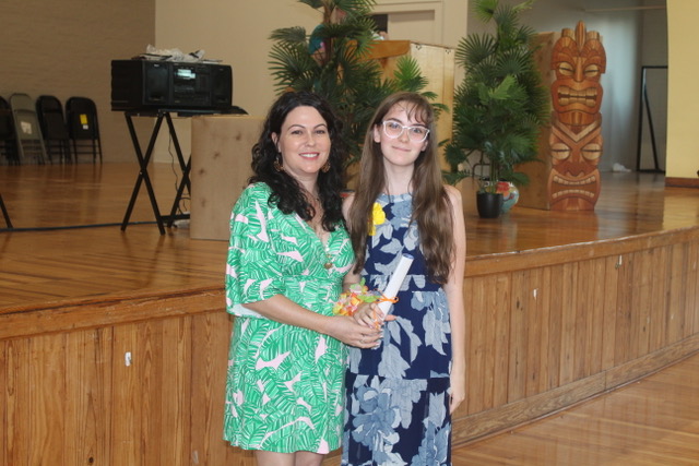 Two people stand together in a decorated hall with tropical plants and tiki statues.