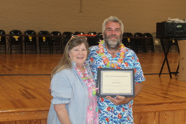 Man and woman smile holding an “Outstanding Supporter” certificate on a wooden stage.