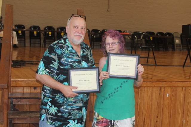 Two people holding “Volunteer of the Year” certificates on a stage with chairs behind them.