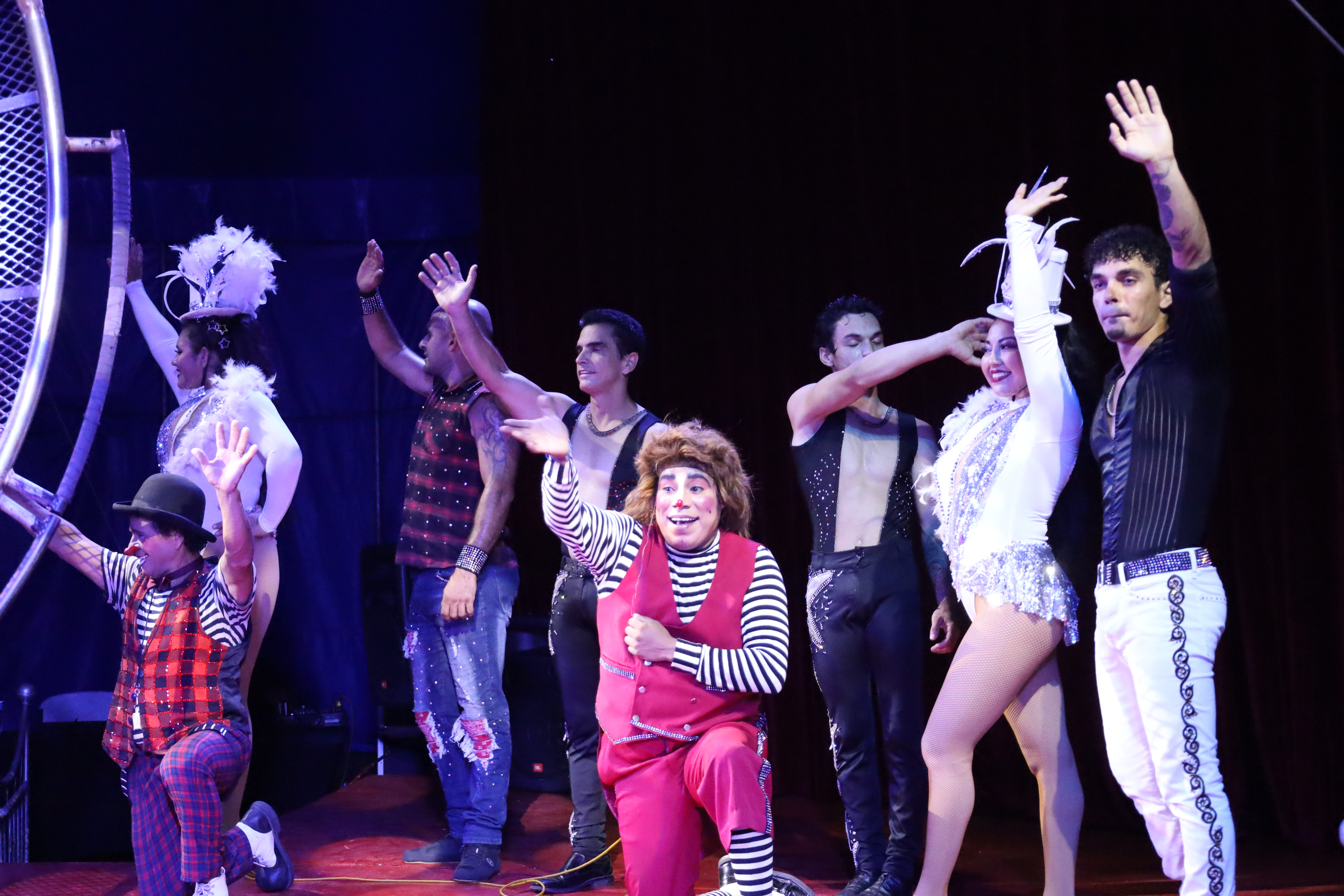 A group of circus performers, including clowns and acrobats in colorful costumes, stand and kneel on stage waving to the audience at the end of a show.