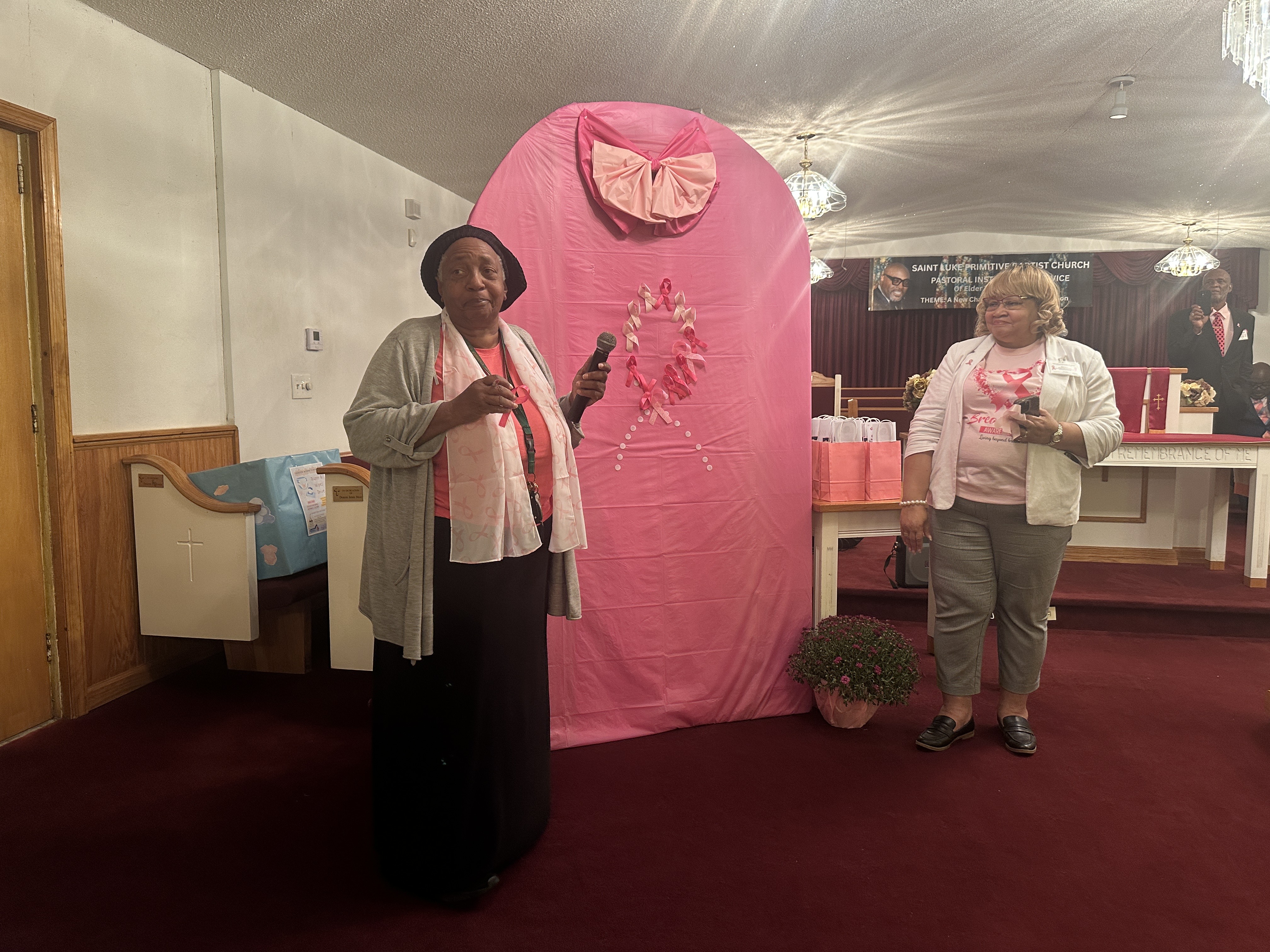 Woman standing at the front of a church speaking into a microphone beside a pink breast cancer awareness display.