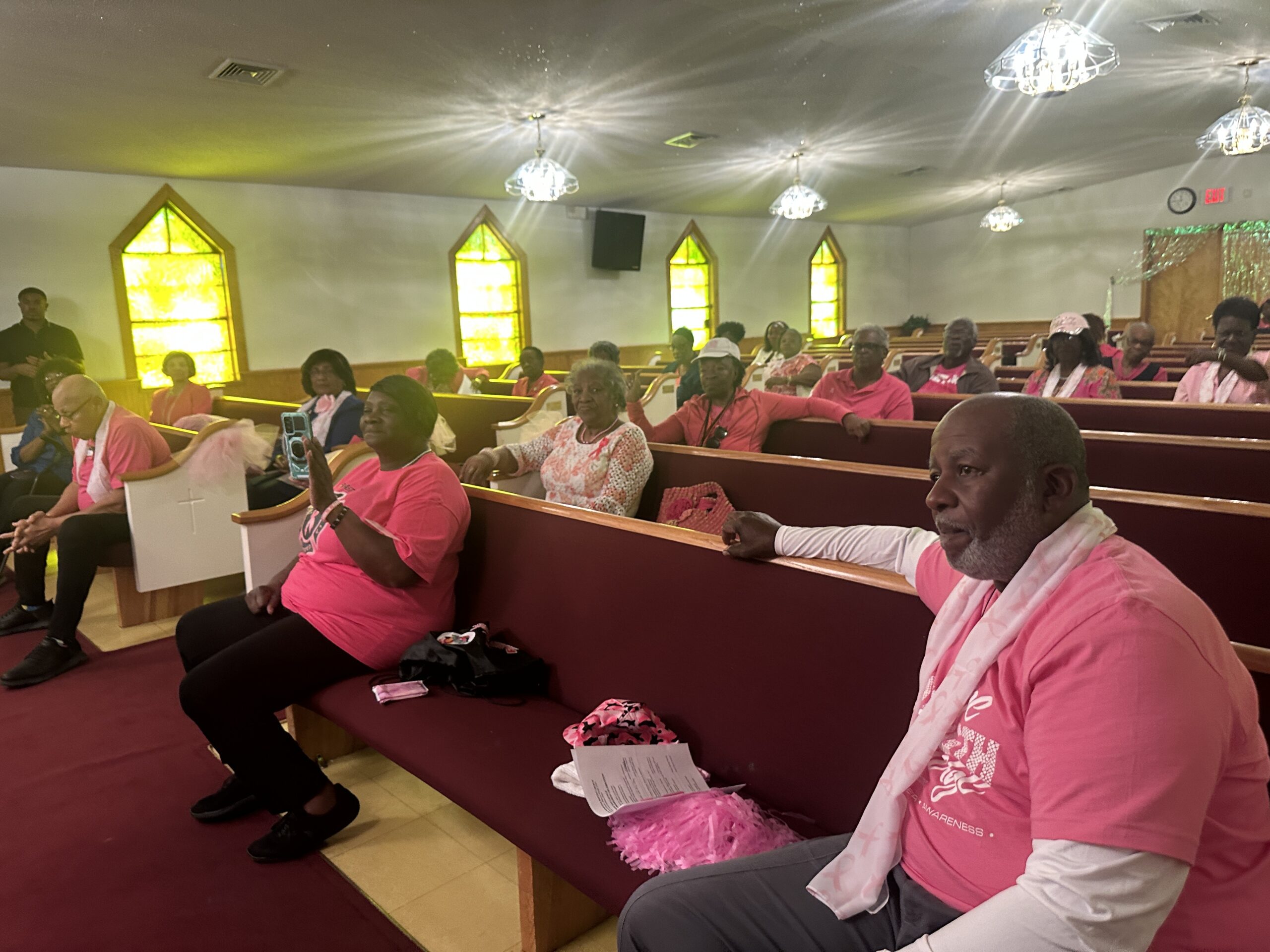 Congregation sitting in church pews wearing pink shirts during a breast cancer awareness service.