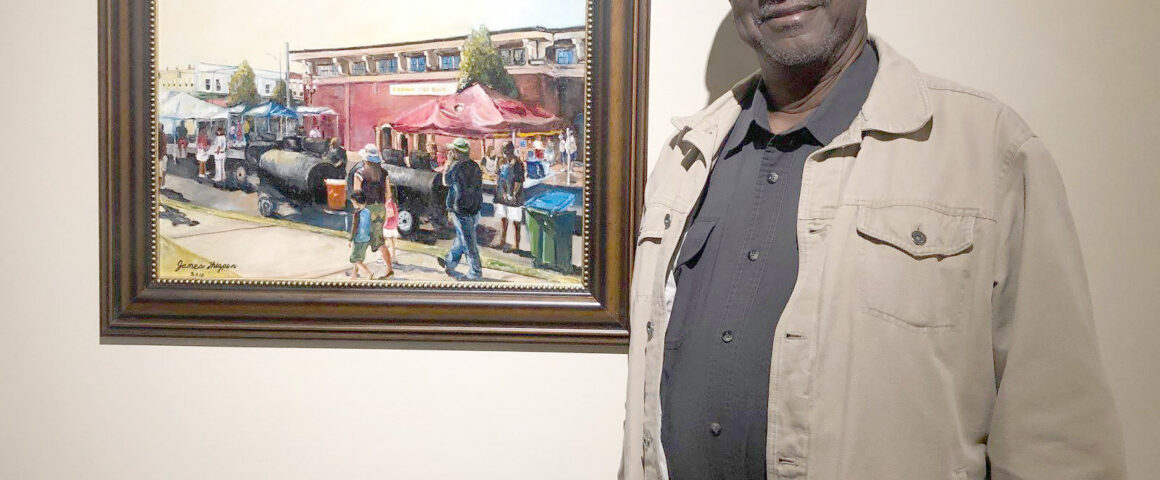 Man standing and smiling next to a framed painting depicting an outdoor market scene with tents and people.