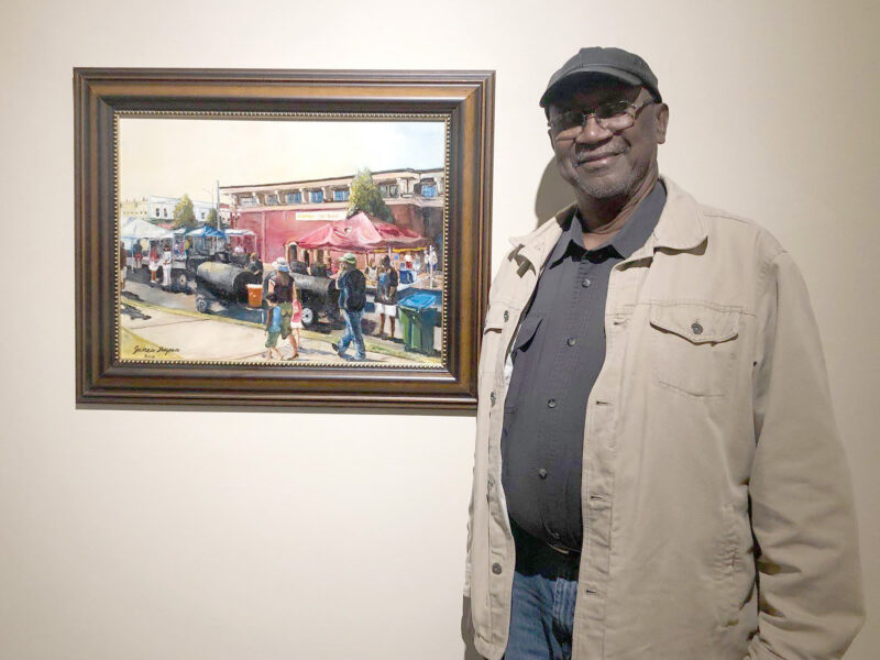 Man standing and smiling next to a framed painting depicting an outdoor market scene with tents and people.