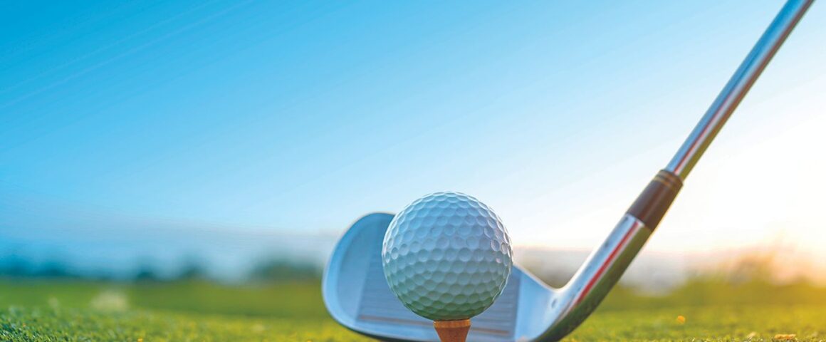 Close-up of a golf club positioned behind a golf ball on a tee at sunrise on a grassy course.