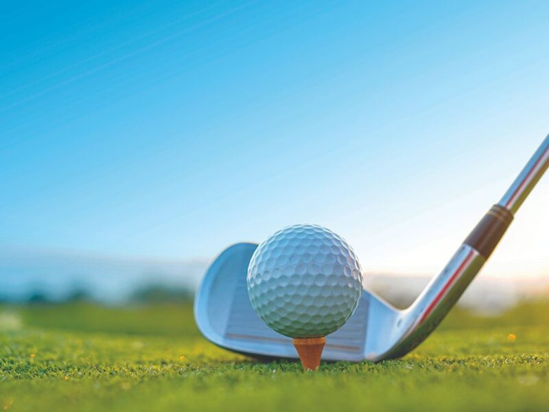 Close-up of a golf club positioned behind a golf ball on a tee at sunrise on a grassy course.