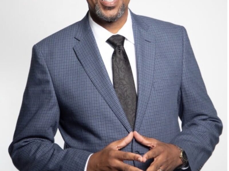 Man in a blue patterned suit jacket, white shirt, and black tie smiling with hands clasped together against a light background.