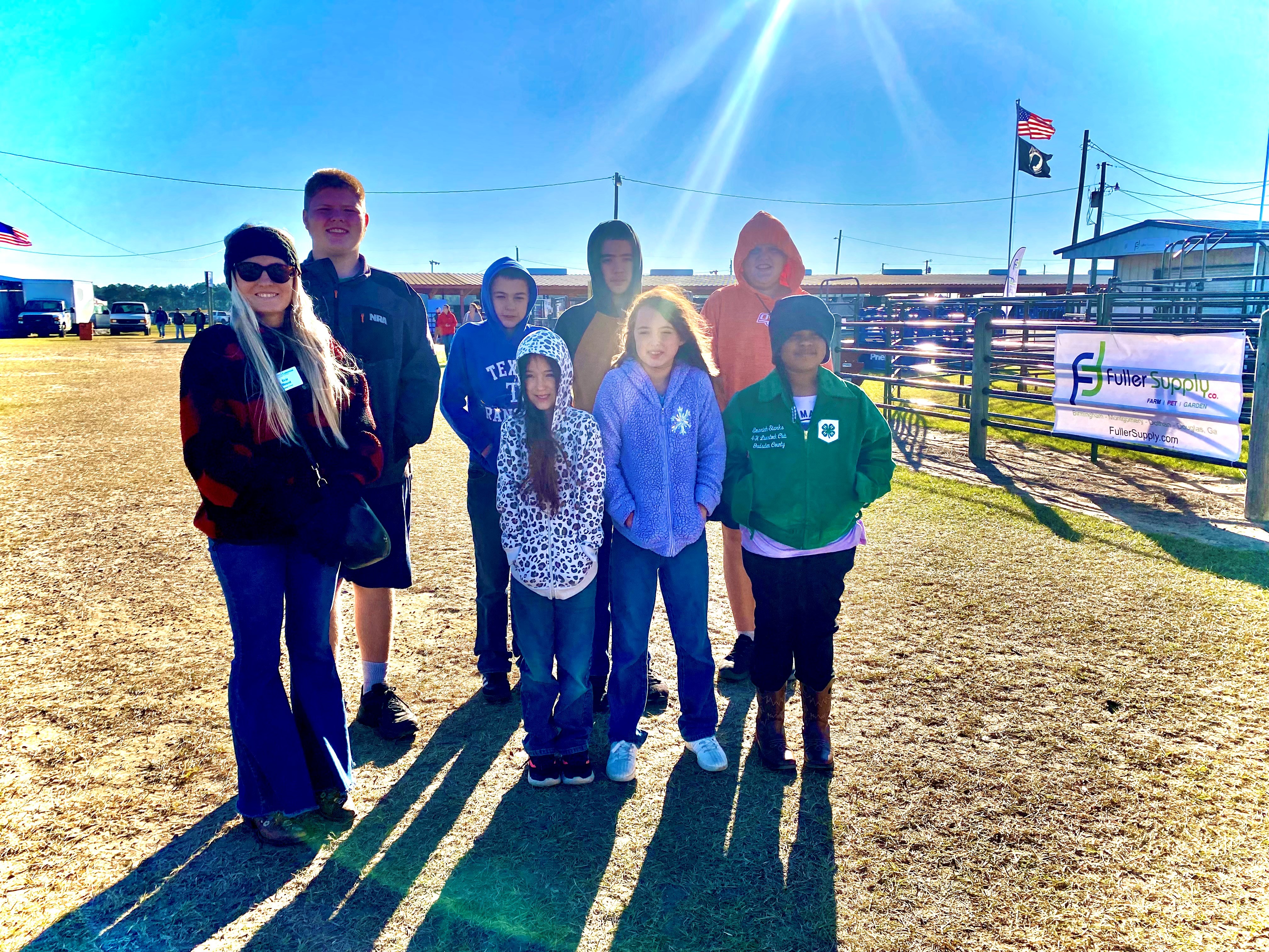 A group of children and one adult stand outdoors on a sunny day in front of a fairground area. They wear jackets, hoodies, and hats, smiling and posing for the photo with shadows cast on the ground.