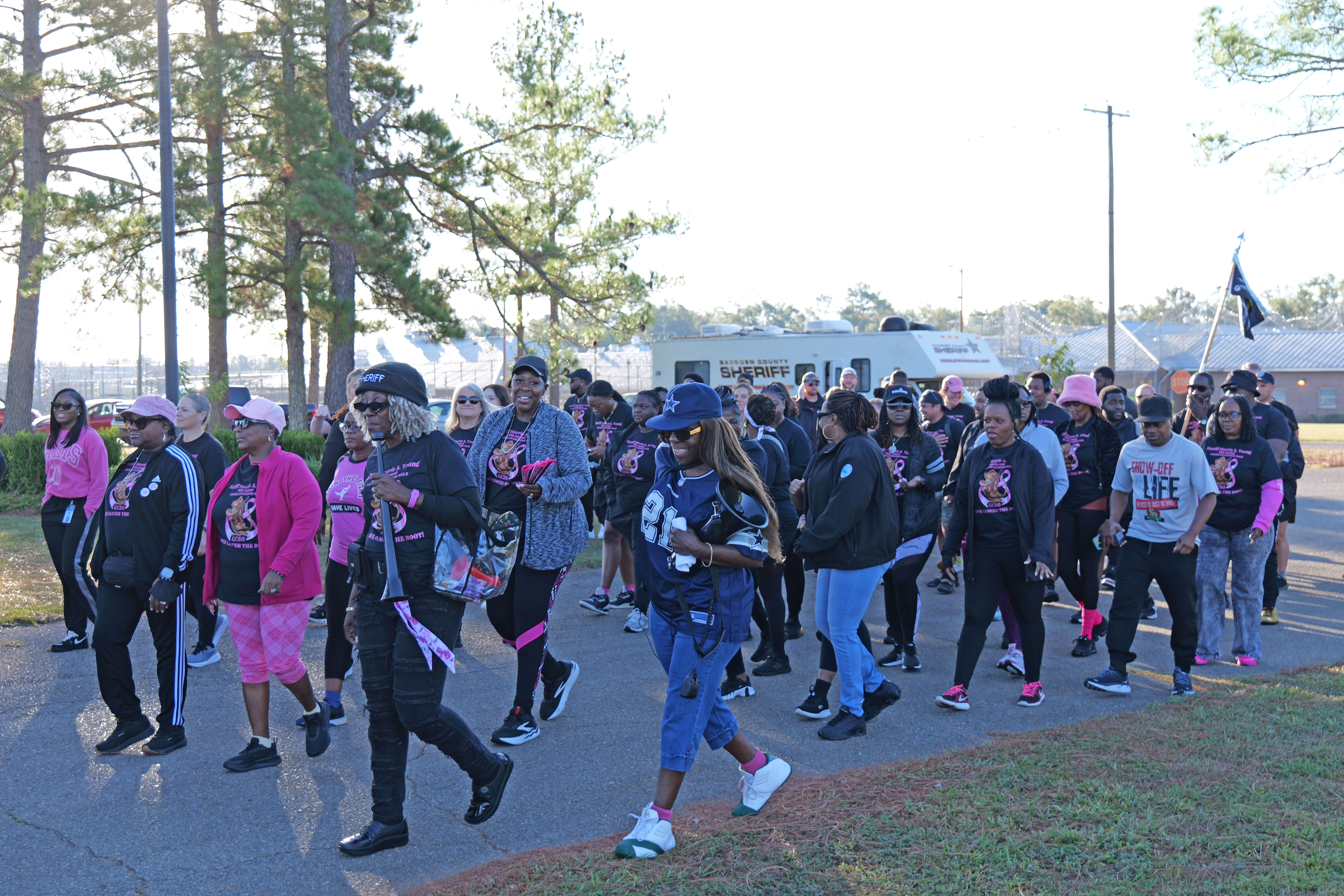 Group photo of participants wearing matching black shirts with pink breast cancer awareness logos, posing together on grass under tall pine trees.