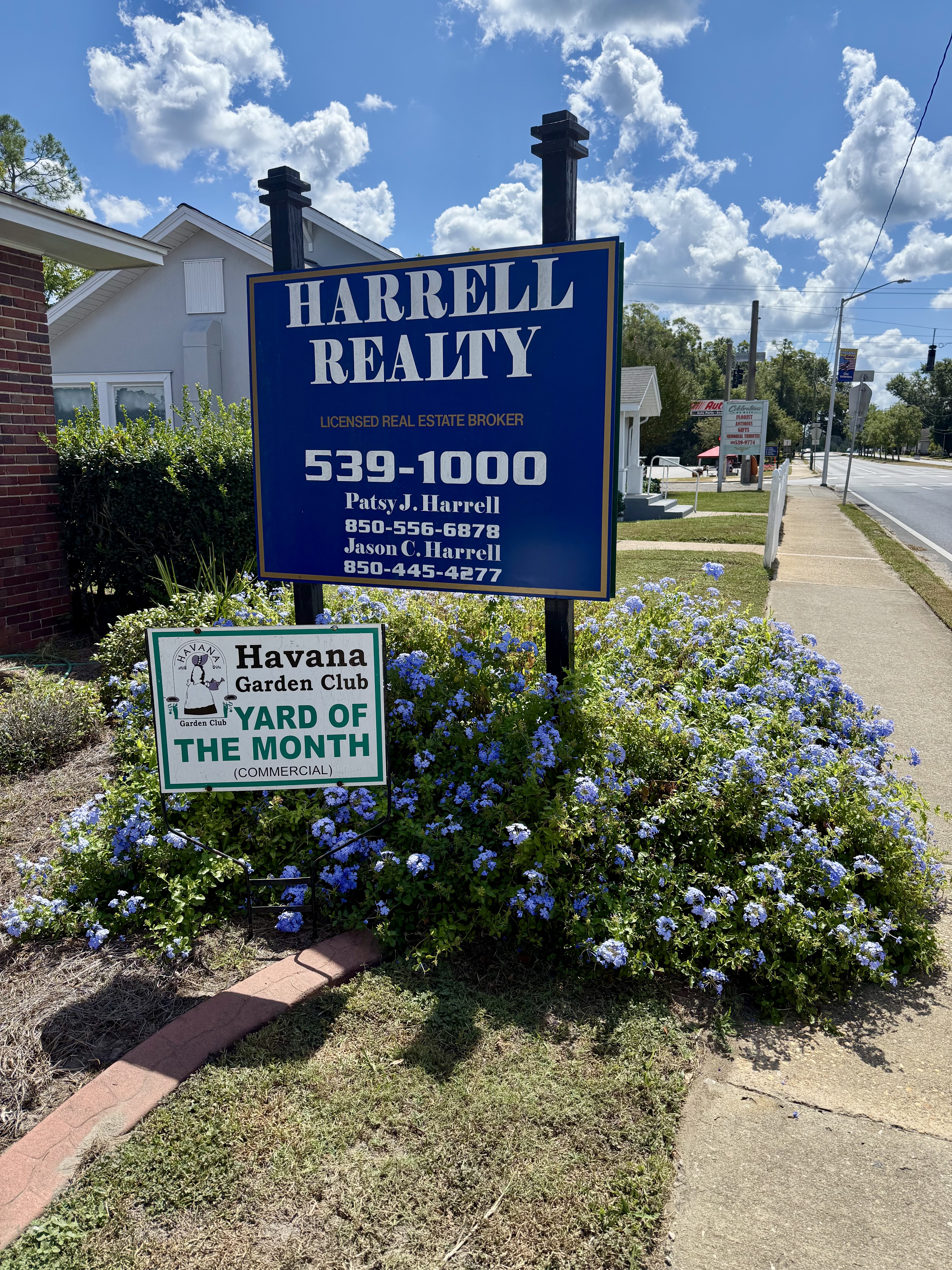 Real estate office sign with flowers and “Yard of the Month” award in front of it.