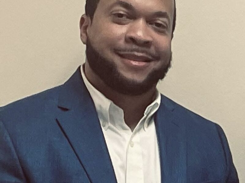 Portrait of a man wearing a blue blazer and white shirt, smiling while standing against a plain indoor background.