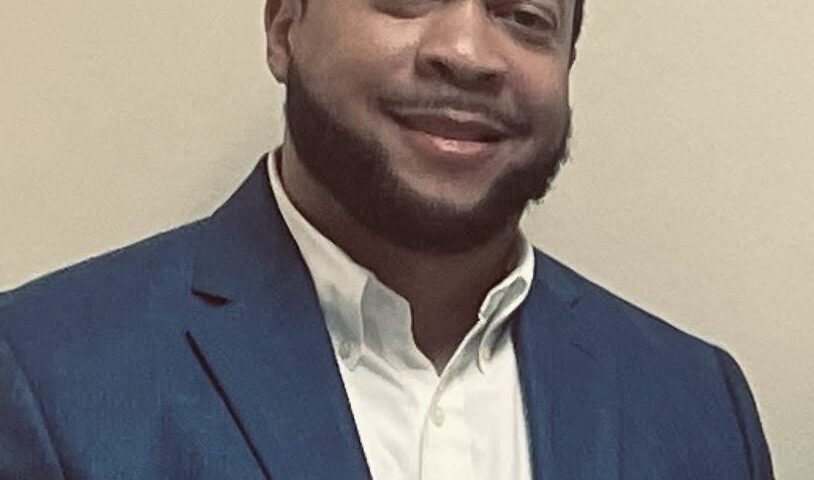 Portrait of a man wearing a blue blazer and white shirt, smiling while standing against a plain indoor background.