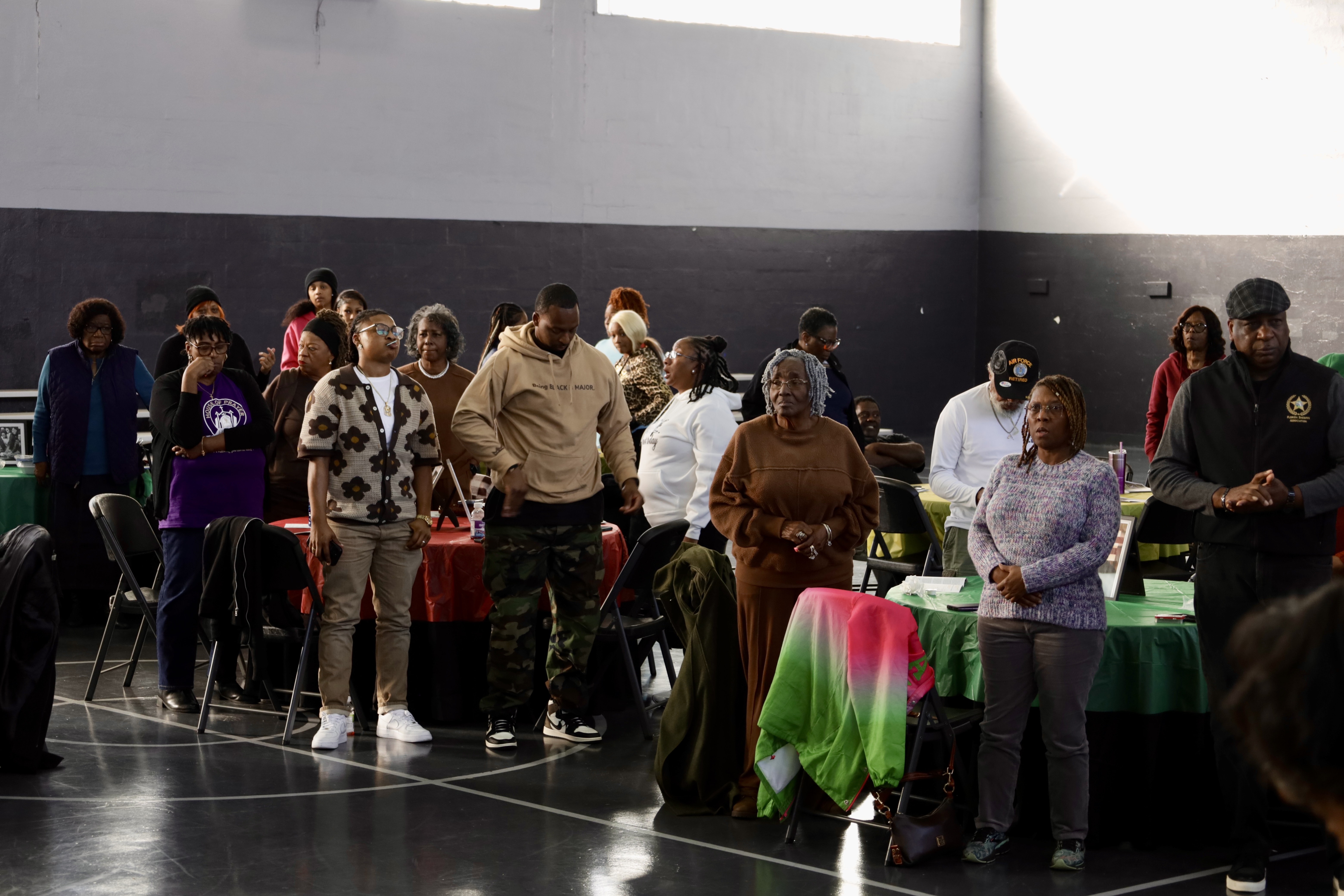 A man standing at a podium indoors, speaking into a microphone during a formal event, with a balloon display in red, black, green, and yellow behind him.