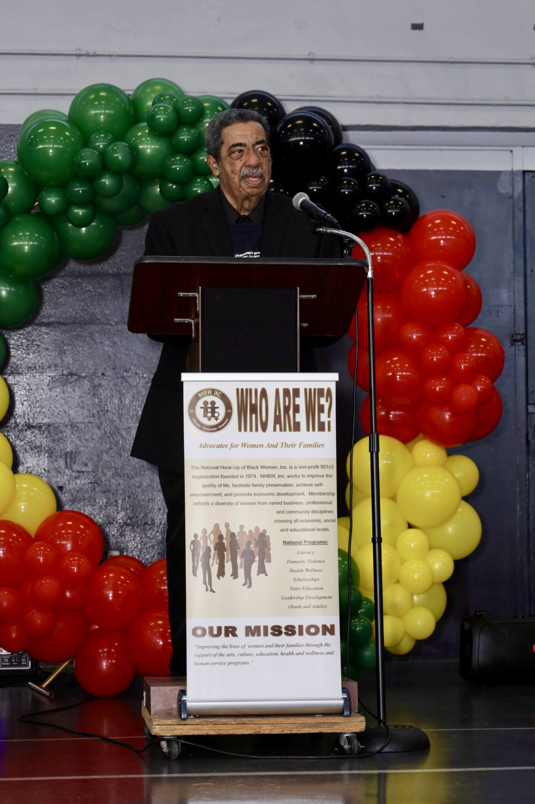 A community meal service scene with volunteers standing behind tables covered in colorful tablecloths, serving food and drinks to attendees inside a gymnasium.