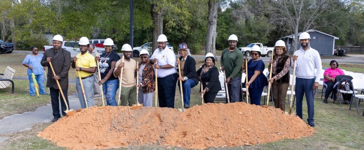 A group of adults wearing white hard hats pose outdoors with ceremonial shovels in front of a mound of dirt, suggesting a groundbreaking ceremony. Trees, parked vehicles, and folding chairs are visible in the background.