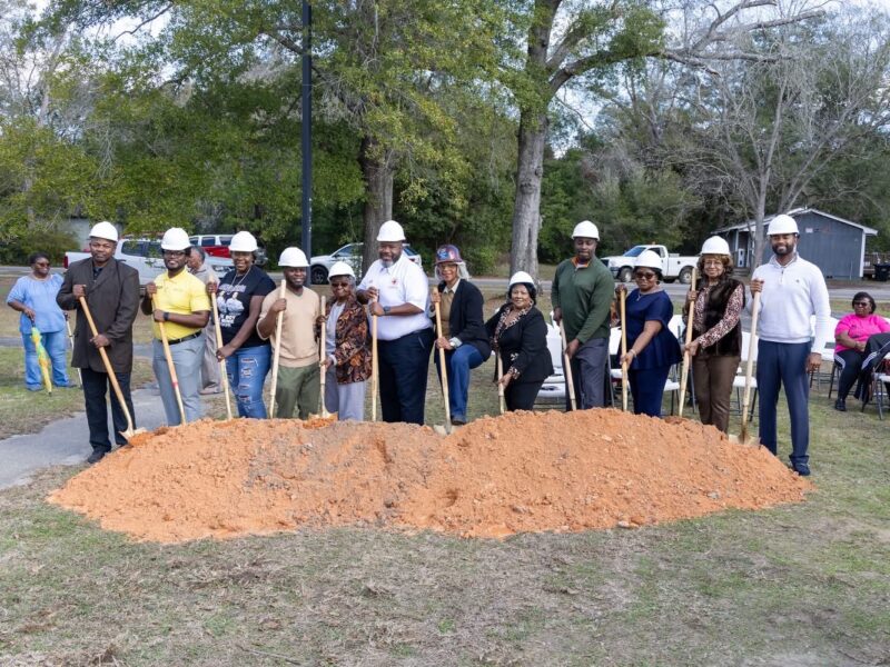 A group of adults wearing white hard hats pose outdoors with ceremonial shovels in front of a mound of dirt, suggesting a groundbreaking ceremony. Trees, parked vehicles, and folding chairs are visible in the background.