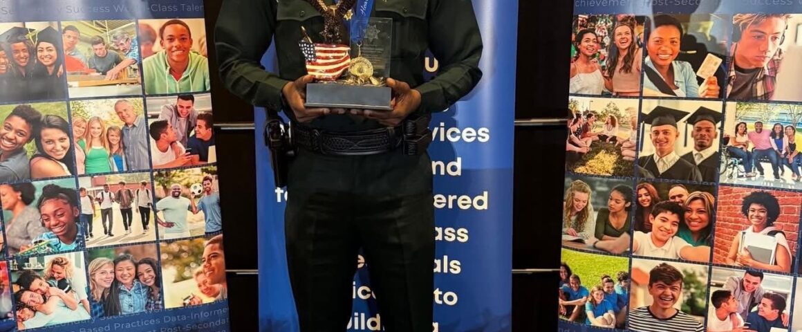 Uniformed law enforcement officer standing indoors between Florida Department of Juvenile Justice banners, holding an eagle statue award mounted on a base with an American flag design.