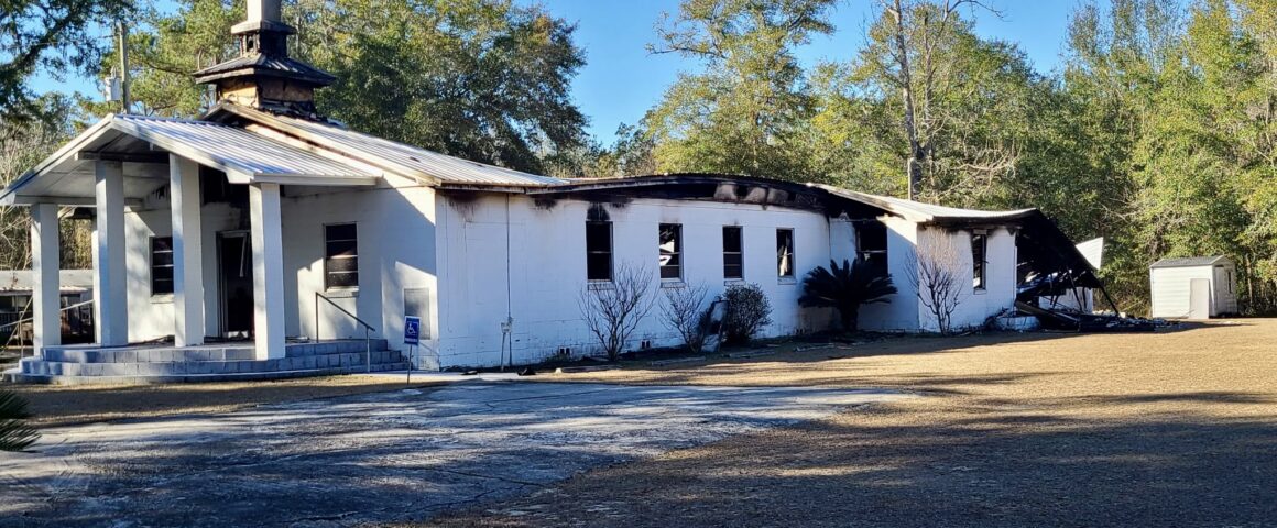 An exterior view of a church building showing significant fire damage to the roof and walls, photographed from a distance in daylight.