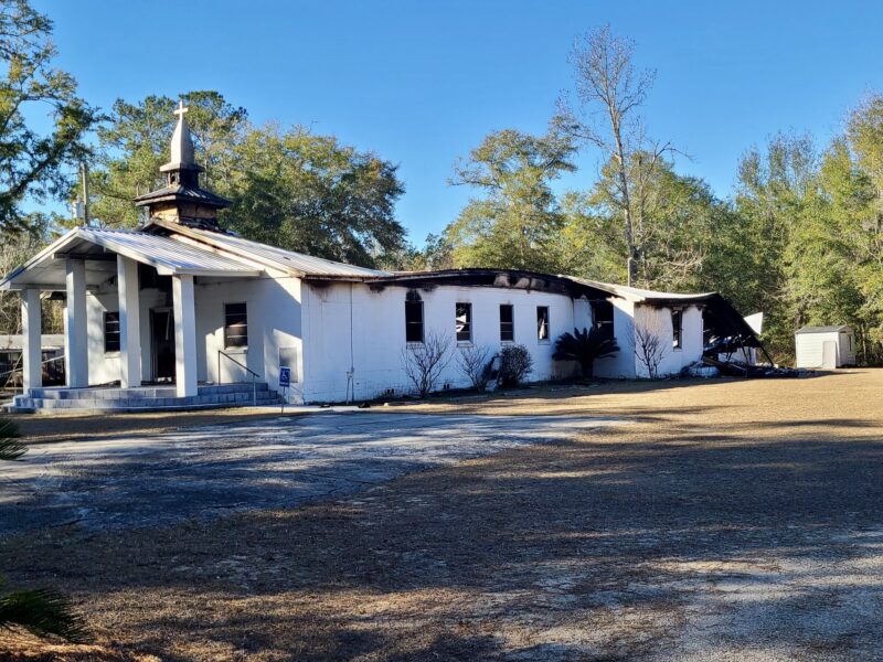 An exterior view of a church building showing significant fire damage to the roof and walls, photographed from a distance in daylight.