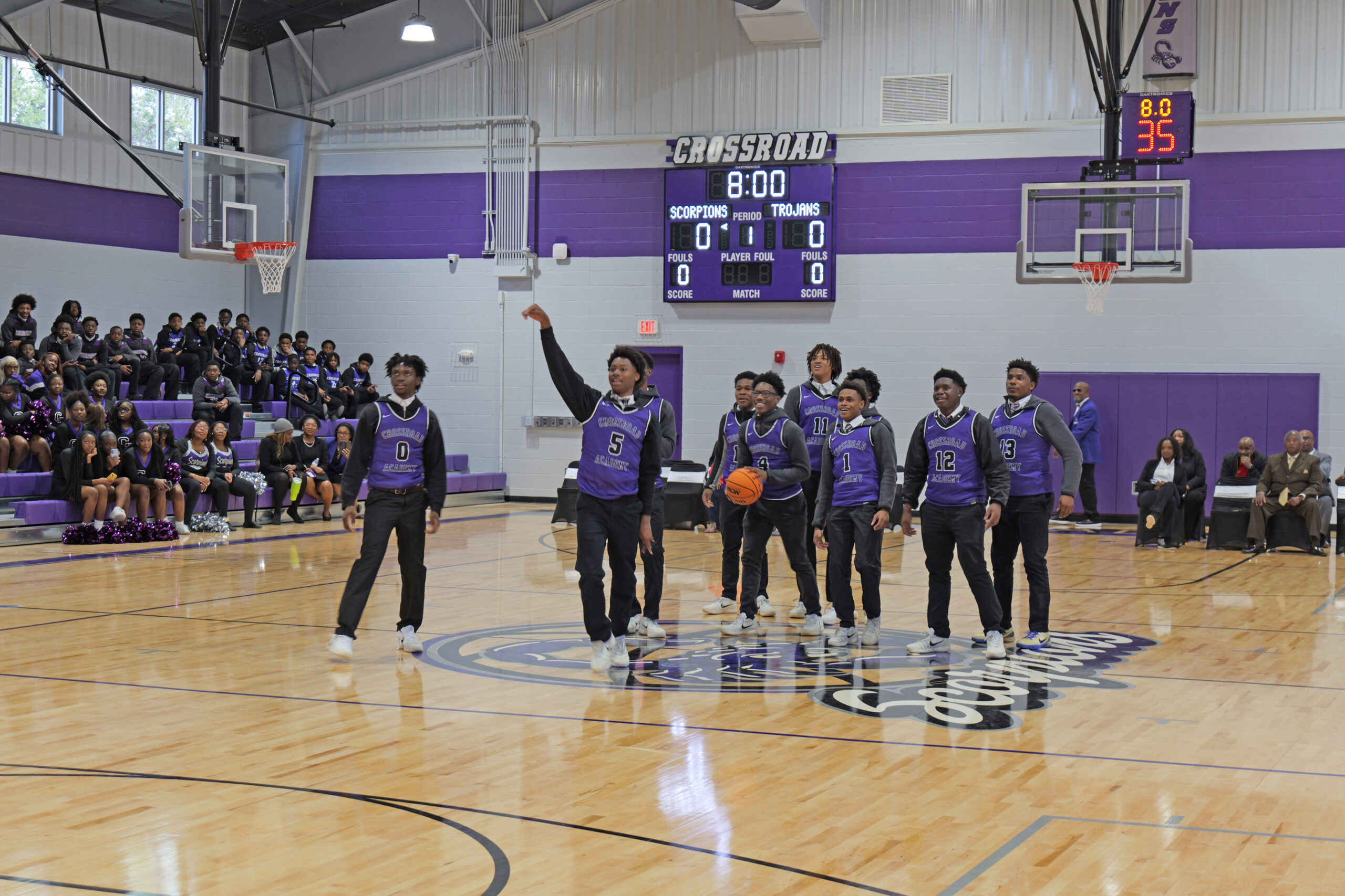 A boys’ basketball team in purple jerseys stands at center court inside a gymnasium. One player has just released a basketball, and spectators fill the bleachers behind them. A scoreboard labeled “Crossroad” is visible above the court.