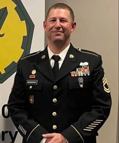 U.S. Army sergeant in formal dress uniform standing indoors, wearing service ribbons and insignia, smiling toward the camera.