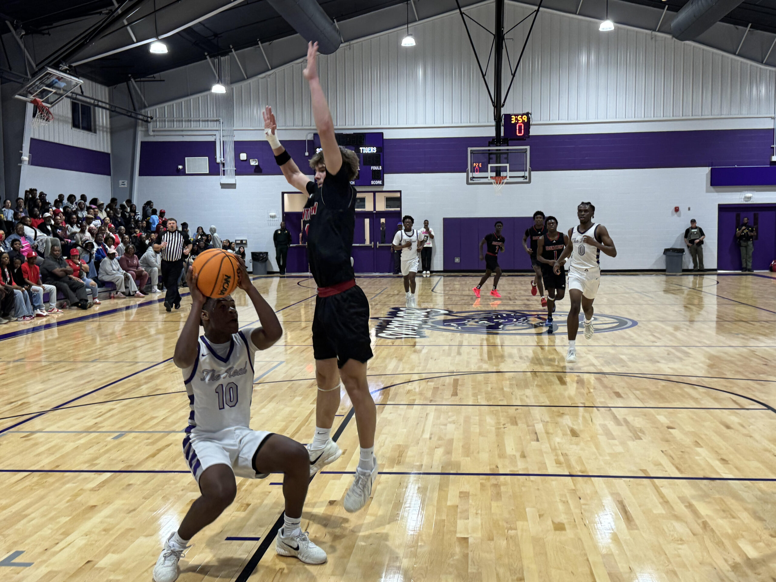 High school basketball game action shot with player attempting a layup while defender contests, packed gym crowd in background, local sports coverage photo.