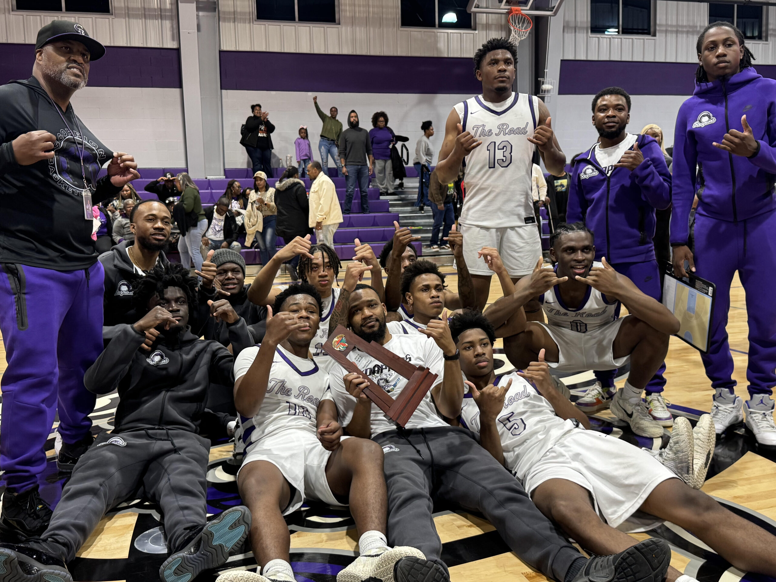 High school basketball players and coaches pose on a gym floor after a game, several holding up hand signs while one player in a white “The Road” jersey holds a wooden plaque; purple bleachers and spectators fill the background.
