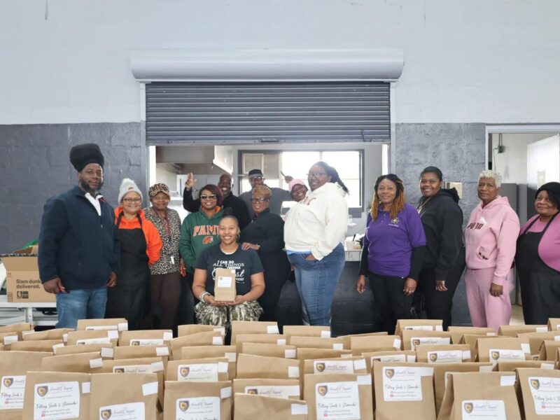 Large group photo indoors showing volunteers standing behind long tables filled with labeled brown paper bags. The group poses and smiles inside a community or gym-like space with a roll-up door in the background.