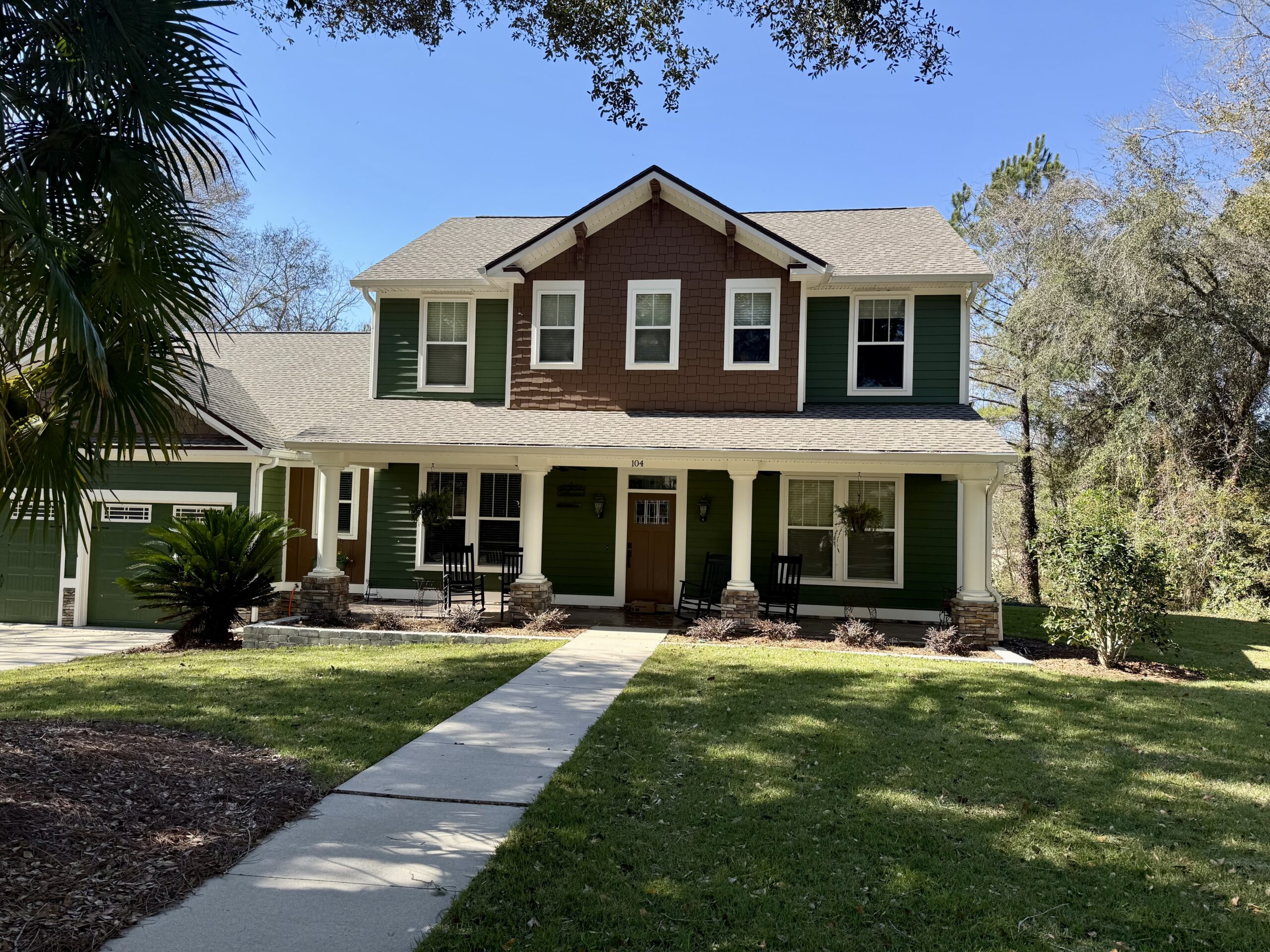 A two-story green house with a covered front porch, white columns, and a central walkway leading through a neatly maintained lawn under a clear blue sky.
