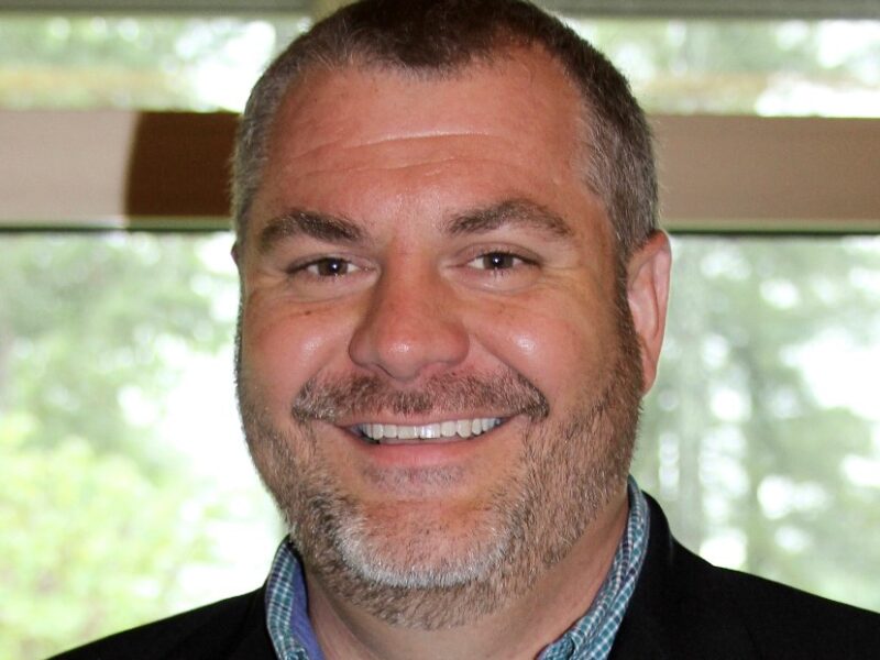 A close-up portrait of a smiling man wearing a dark blazer over a collared shirt. He is photographed indoors with soft lighting and a blurred background, giving the image a professional headshot look.