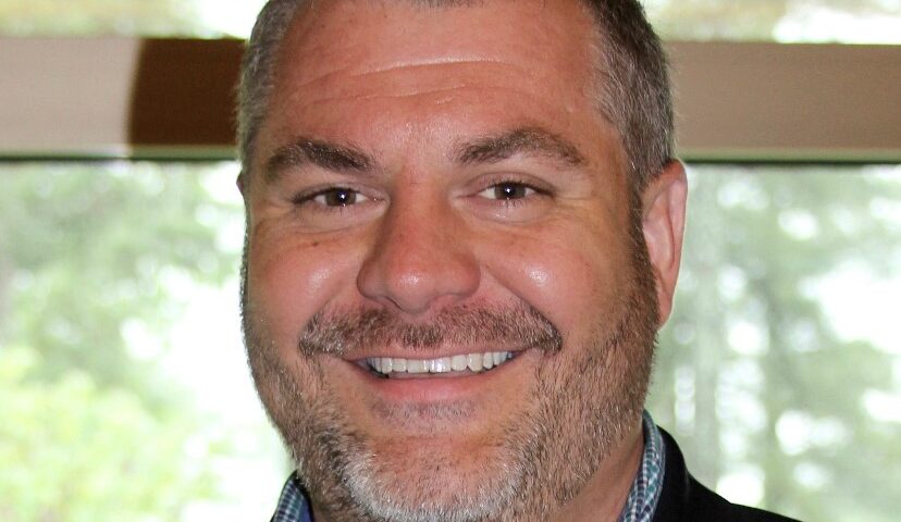 A close-up portrait of a smiling man wearing a dark blazer over a collared shirt. He is photographed indoors with soft lighting and a blurred background, giving the image a professional headshot look.