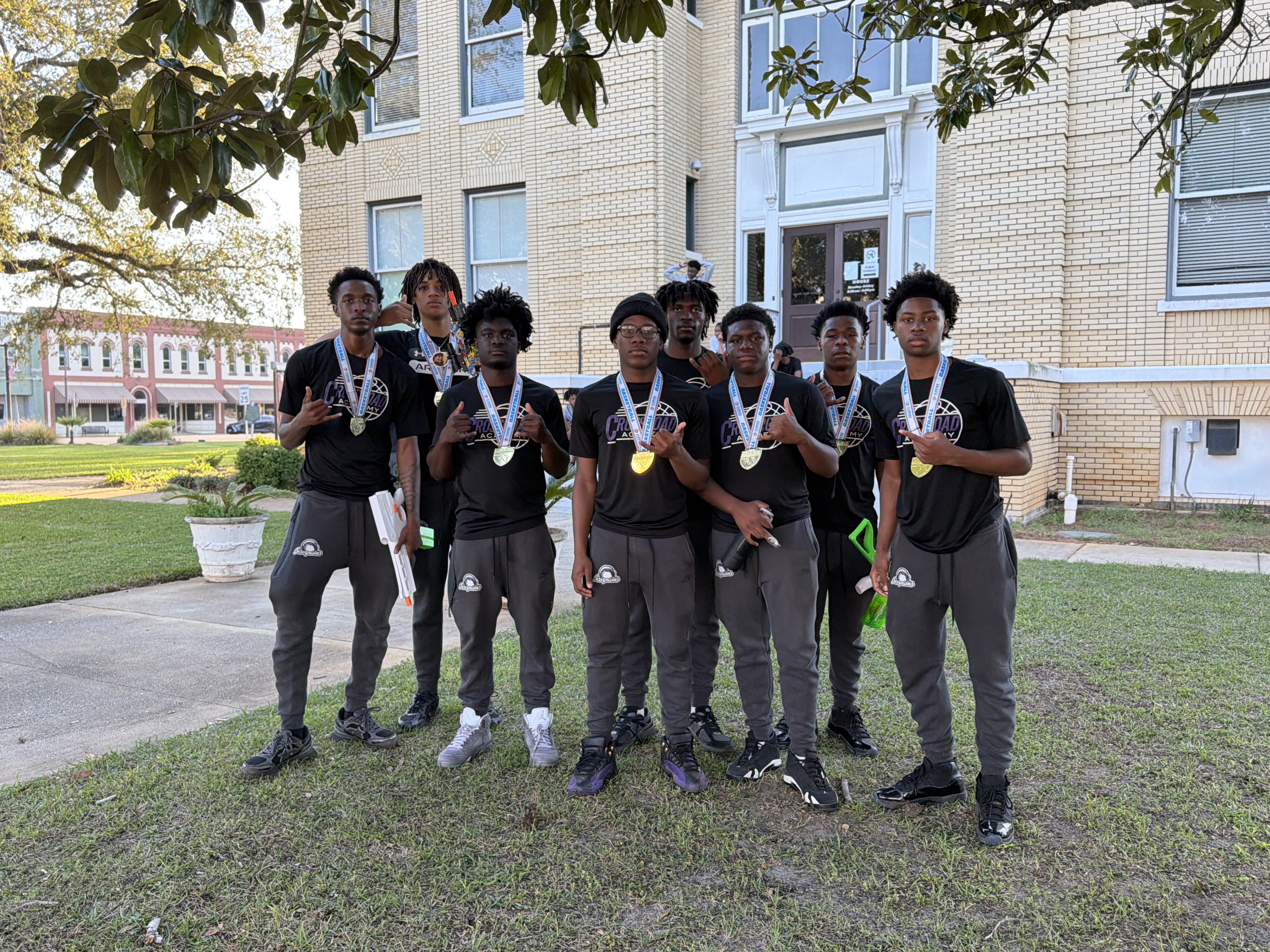 Group of young athletes wearing medals and matching black outfits posing in front of a historic building after a community sports event.