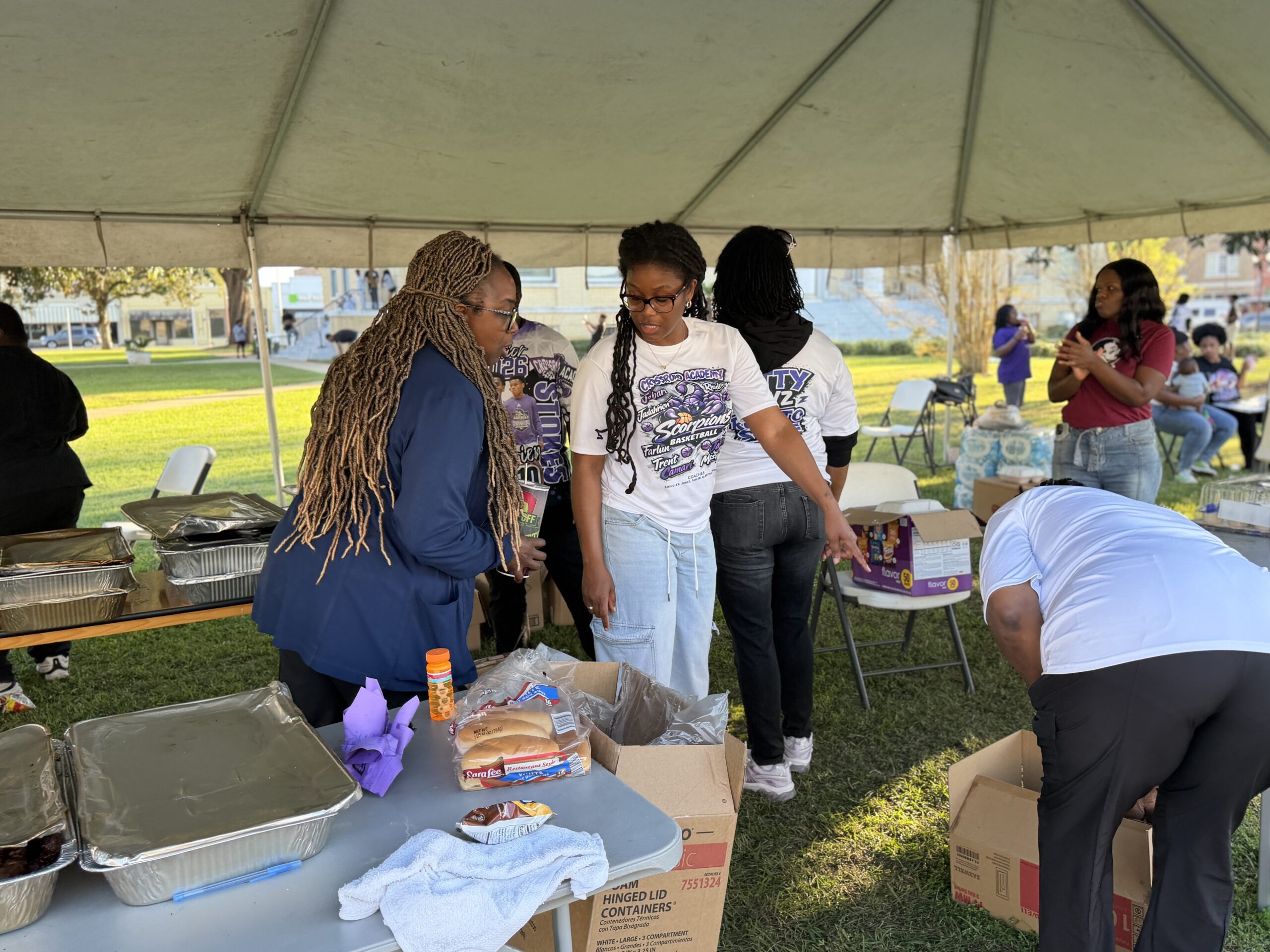 Volunteers preparing and organizing food under a tent at an outdoor community gathering with tables of supplies and serving trays.