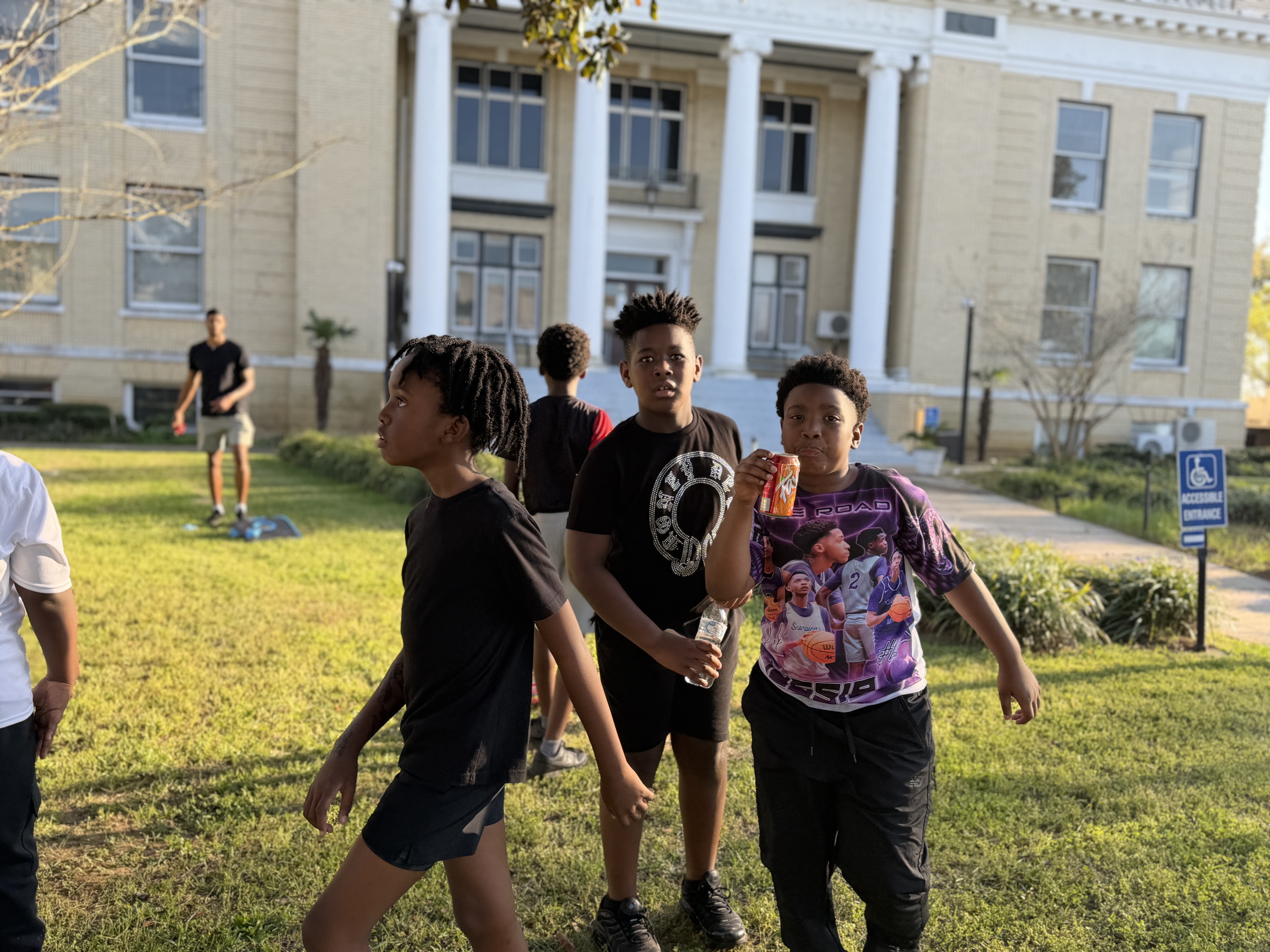 Children walking across a grassy area in front of a historic building during a community event, with one child drinking from a soda can.